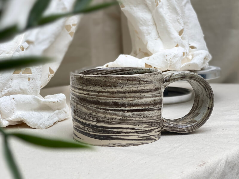 A grey and white marbled pottery mug sits on a table covered in beige fabric